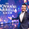 Giovanni Zarrella grinst und zeigt mit ausgestrecktem Arm auf eine Leinwand mit dem Logo der Giovanni Zarrella Show