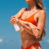 Frau im Bikini am Strand cremt sich mit Sonnencreme ein vor blauem Himmel