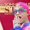 Julian F. M. Stoeckel guckt überrascht mit Hand vor dem Mund vor dem Logo von "Das Sommerhaus der Stars"