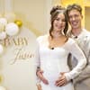 Valentin Lusin steht hinter Renata Lusin in einem weißen Kleid und umarmt sie bei ihrer Babyparty