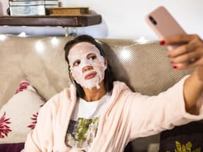 Frau sitzt mit Tuchmaske auf Bett und macht Selfie