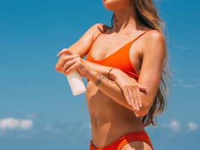 Frau im Bikini am Strand cremt sich mit Sonnencreme ein vor blauem Himmel