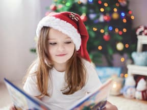 Mädchen mit Weihnachtsmütze sitzt vor einem Tannenbaum und ließt ein Buch