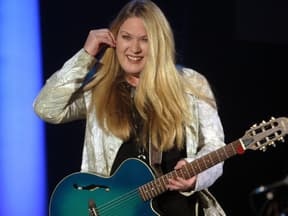 Juliane Wedding steht mit Gitarre auf der Bühne und fasst sich lächelnd ans Ohr