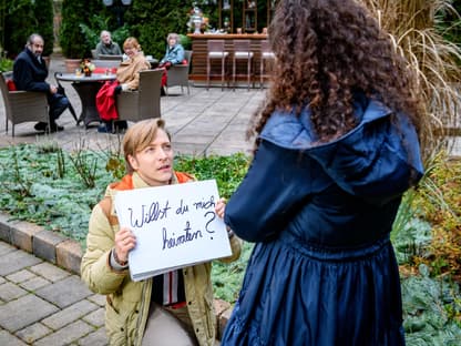 Sturm der Liebe Gerry kniet vor Shirin auf dem Boden und hält ein Schild mit den Worten "Willst du mich heiraten?" vor sich