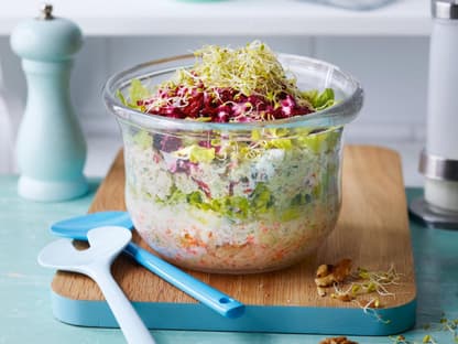 Schichtsalat mit Feta-Dressing in einer Glasschale auf einem Holzbrett mit blauem Besteck daneben