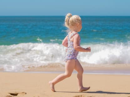 Blondes Mädchen rennt am Strand vor dem Meer rum und trägt einen Badeanzug