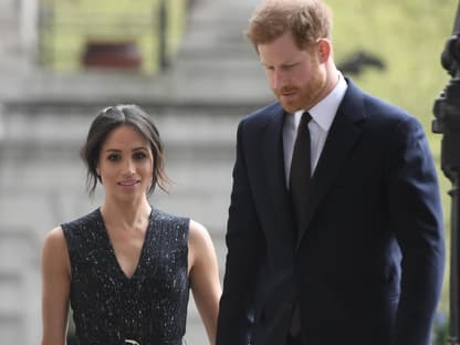 Prinz Harry sieht zu Boden, Herzogin Meghan läuft hinter ihm