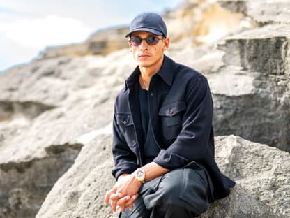 Emilio Sakraya mit Sonnenbrille auf sitzt auf einem Stein am Strand